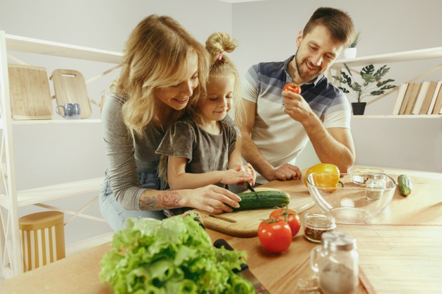 Cute little girl and her beautiful parents are cutting vegetables and smiling