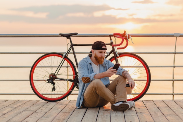 man wearing denim shirt and cap holding smartphone with bicycle in morning 