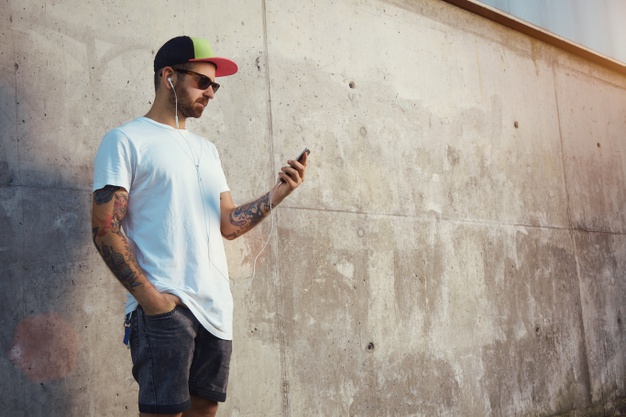 Young man standing next to a gray concrete wall looking at his smartphone's screen and listening to music in his white earplugs 