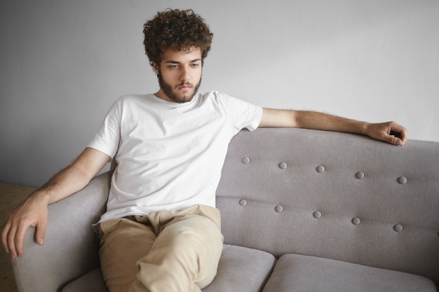 young caucasian man with voluminous hair and thick beard sitting casually on gray sofa at home