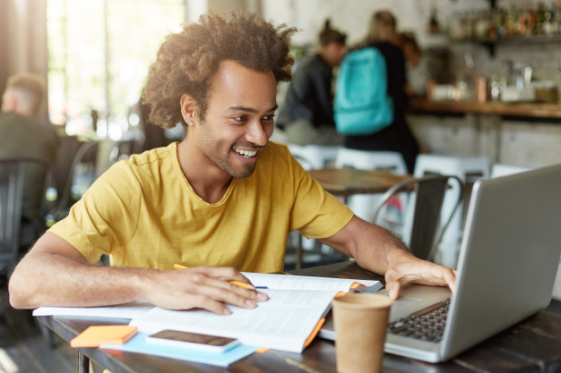  happy student male with curly hair dressed casually sitting in cafeteria working with modern technologies