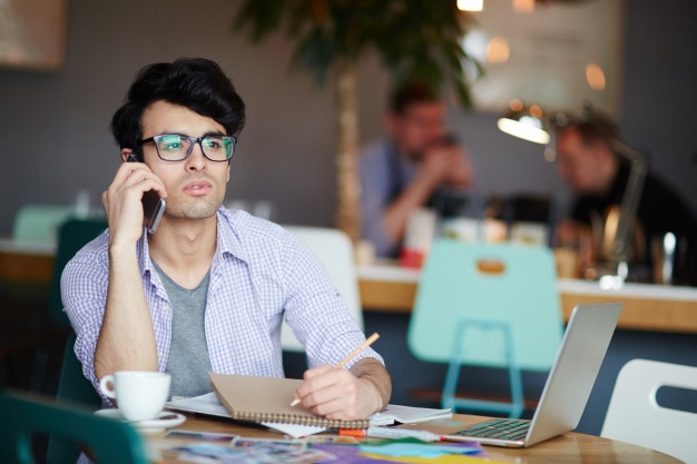 man calling by phone while making notes and working in cafe