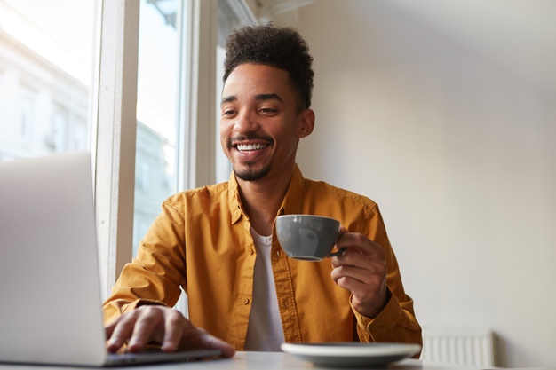 american sitting at table in cafe and working laptop, wears in yellow shirt, drinks aromatic coffee