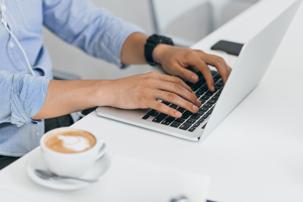 Man in blue shirt using laptop for work, typing on keyboard. indoor portrait of male hands on computer and cup of coffee on table