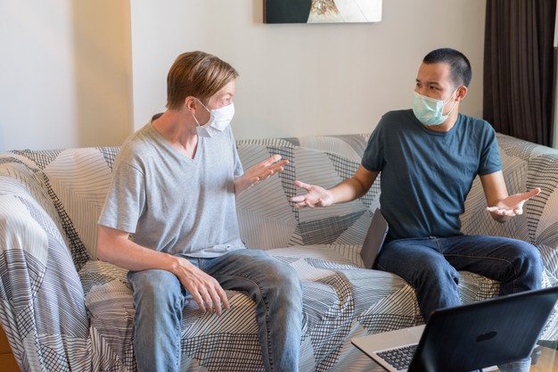 Two confused multi ethnic men with mask as friends shrugging shoulders at home under quarantine