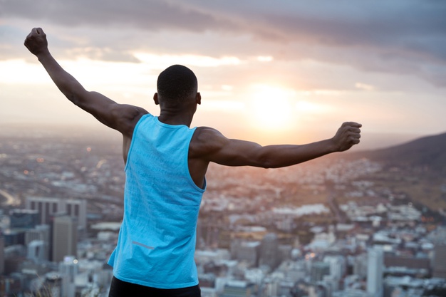 carefree-independent-man-with-dark-skin-stands-back-stretches-hands-as-holding-world-feels-freedom-wears-blue-casual-vest-enjoys-panoramic-view-with-sunrise-recreation-concept