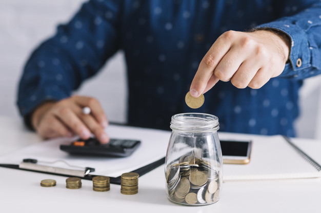 man's hand putting coin in jar using calculator 