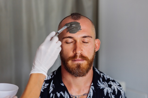 Cosmetologist apply clay mask with brush on young handsome mans face in beauty clinic