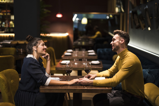 Young smiling man and cheerful woman holding hands at table with glasses of wine in restaurant 