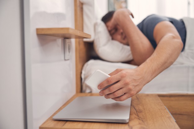 Brunette male staying in bed while looking on screen of mobile and checking time