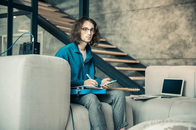 Creating new song. serious inspired guy in clear glasses being involved in creation process while writing down notes