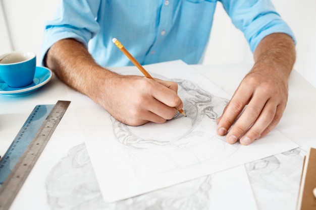Hands of young businessman sitting at table with pencil drawing portrait. white modern office interior