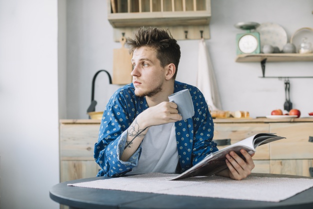 Man looking away holding coffee cup and book at home