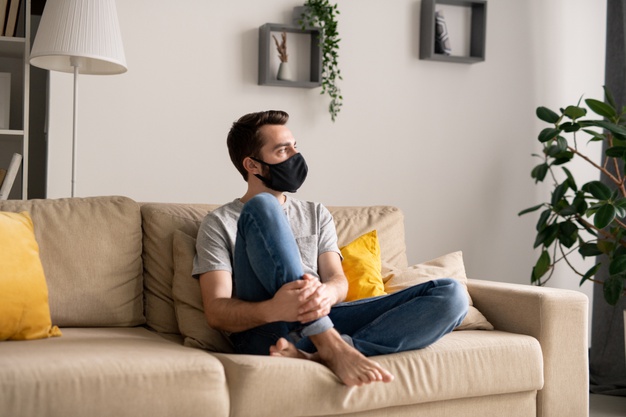 Pensive young man in cloth mask being isolated at home sitting on sofa and looking away