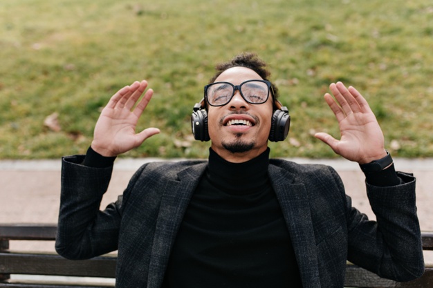 man spending morning outdoor and listening music. photo of handsome african guy in black suit sitting on bench in front of green lawn
