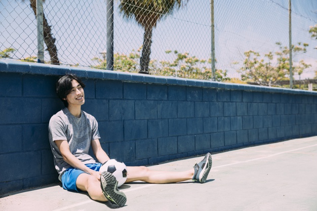 Smiling young man sitting at fence with football