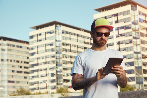 young man wearing white plain t-shirt and red, yellow and black trucker hat looking at his tablet against city buildings and sky