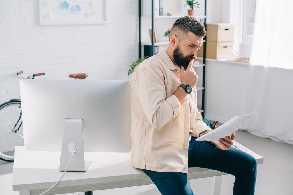 Bearded office worker watching at papers and thinking in modern office 