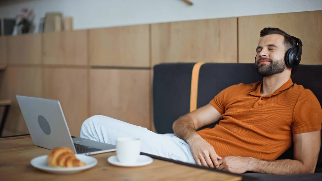 Cheerful man listening music in headphones near breakfast and laptop in cafe