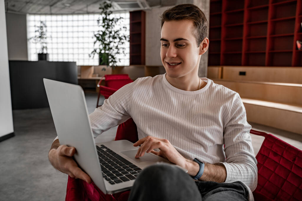 Cheerful young student using laptop while sitting in armchair