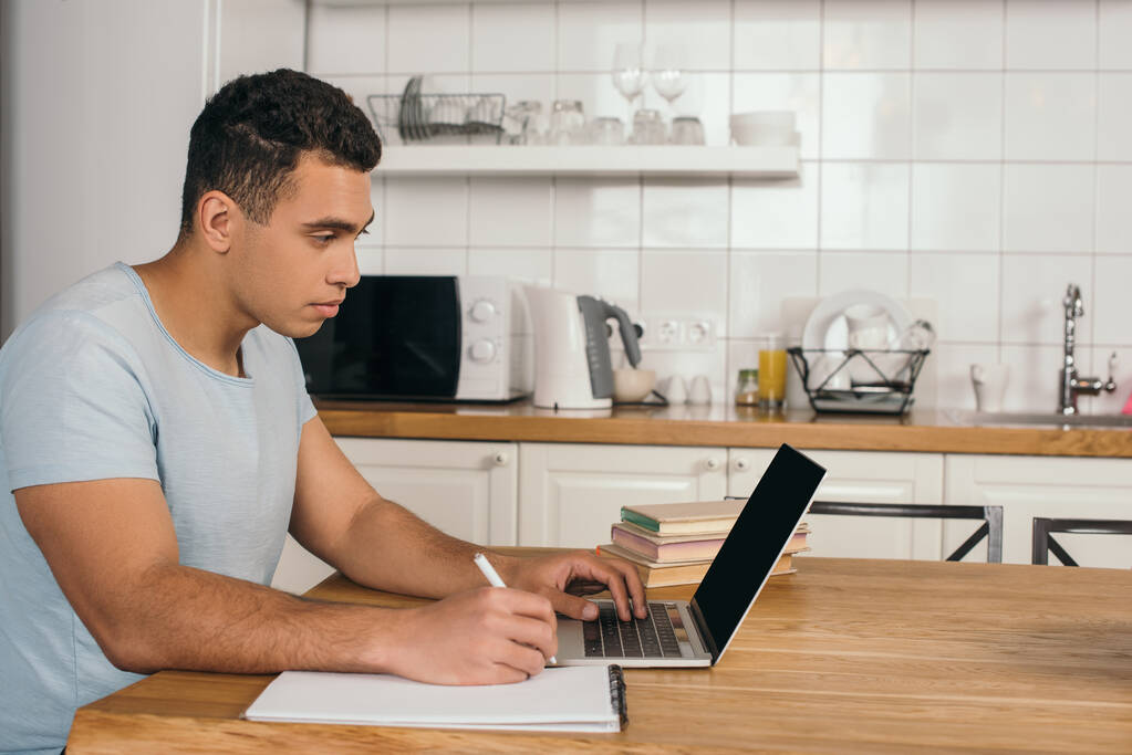 Handsome mixed race man holding pen near notebook and using laptop with blank screen