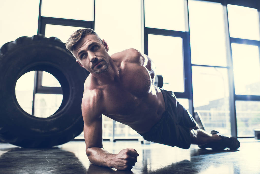 Handsome shirtless sportsman doing plank on one hand in gym