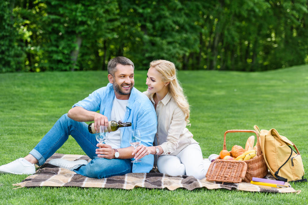 couple drinking wine while sitting together on plaid at picnic