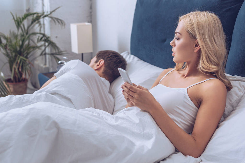 Jealous young woman using smartphone and looking at boyfriend sleeping in bed