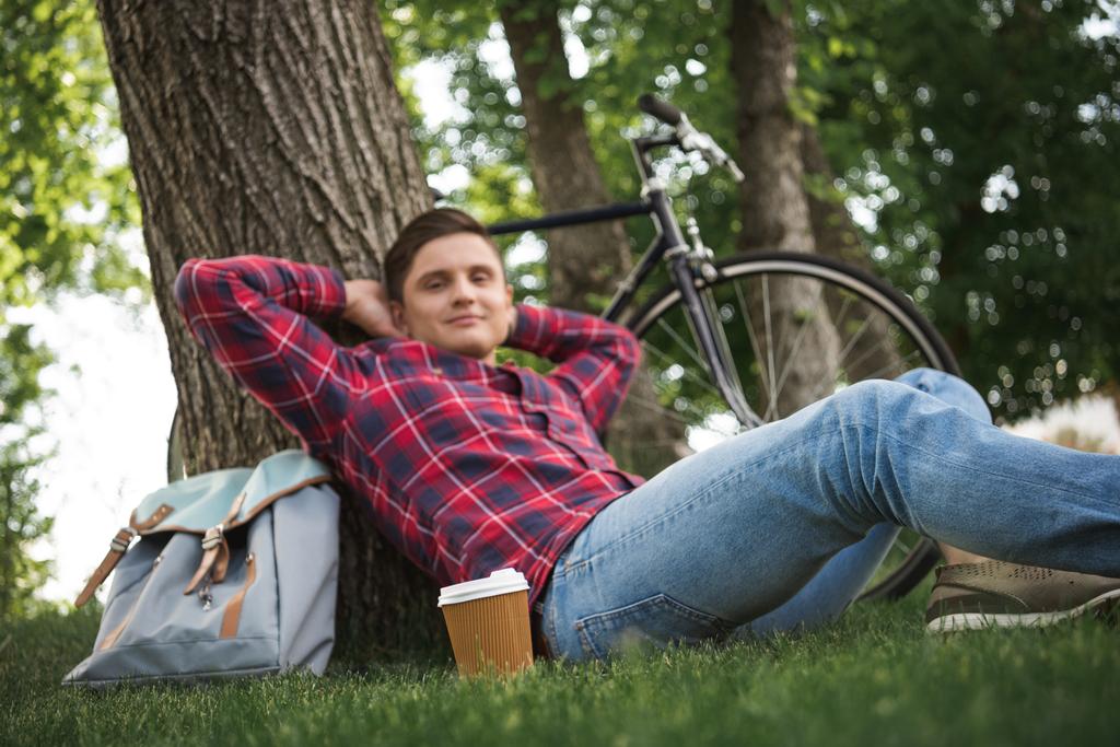 young man leaning to tree trunk with coffee cup and backpack