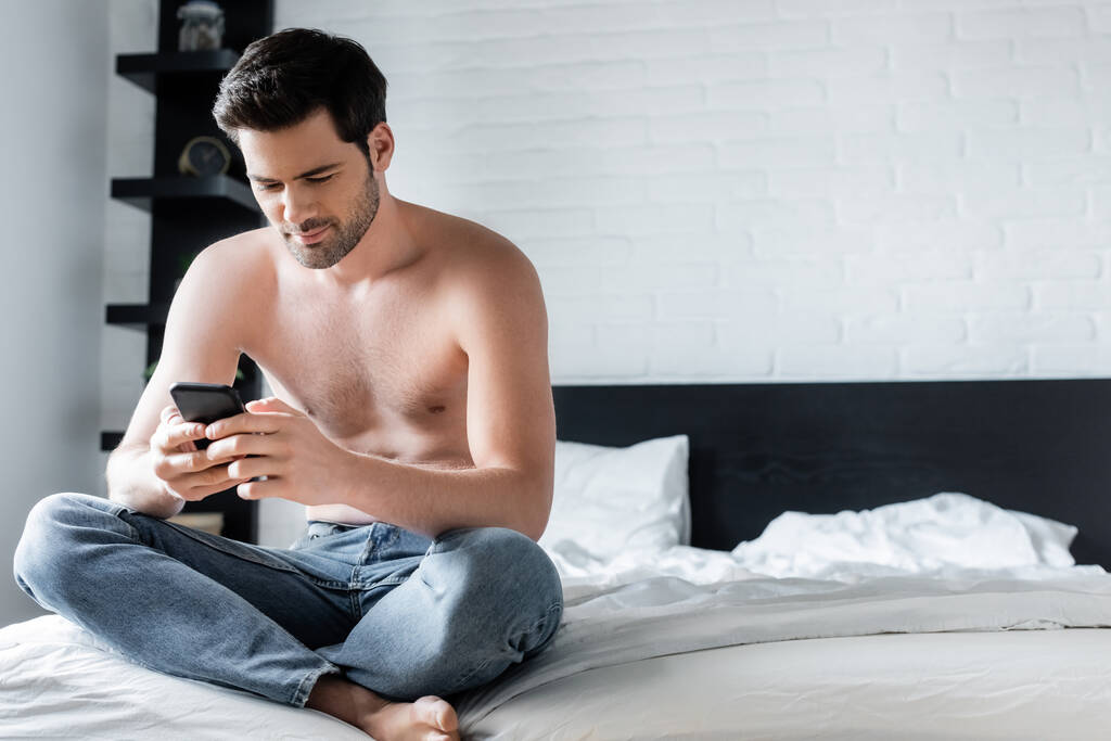 Positive shirtless man using smartphone in bedroom at home