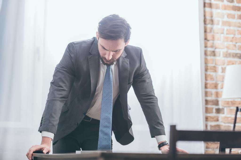 Sad man in gray suit standing near table and looking at photo in frame at home, grieving disorder concept