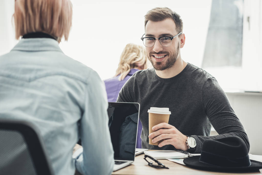 Selective focus of smiling businessman in eyeglasses with coffee to go at workplace in office