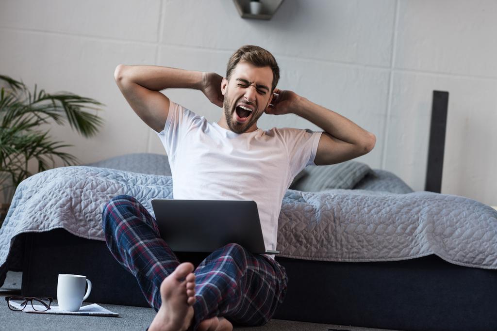  Young man yawning while sitting on floor with laptop in his bedroom 