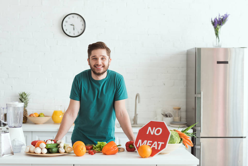 Smiling handsome vegan man standing near kitchen counter with vegetables and no meat sign