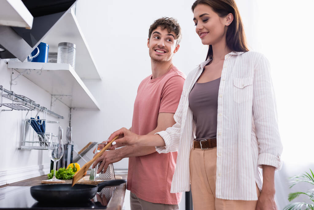 Smiling man looking at girlfriend cooking on stove in kitchen