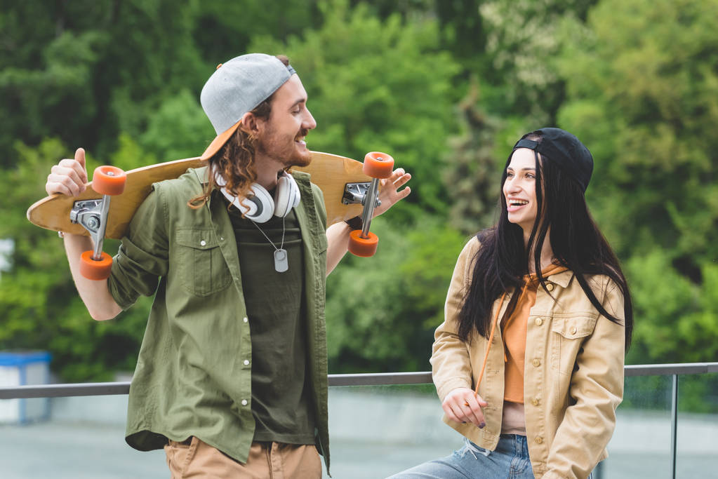 Smiling man with skateboard looking at brunette woman, standing on green trees background