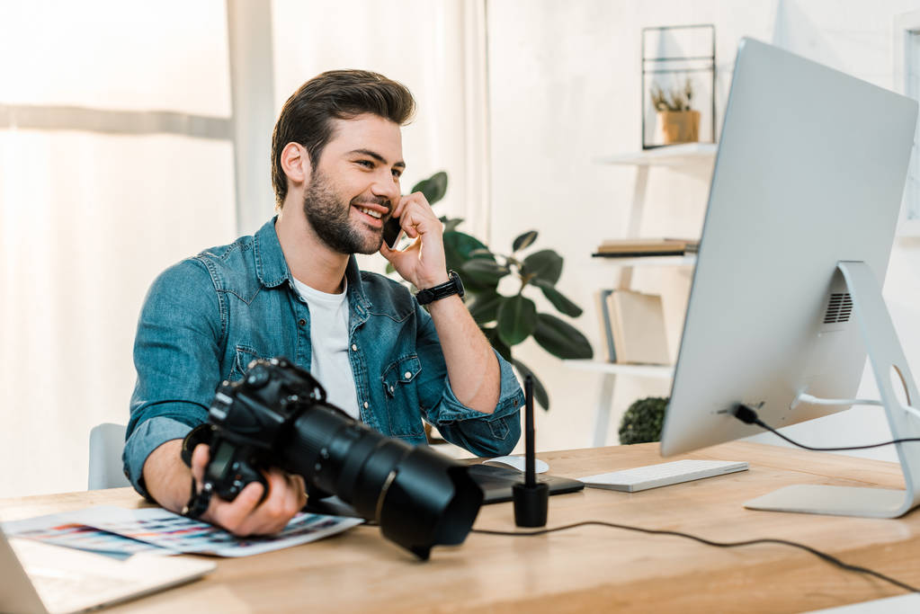 Smiling young photographer holding camera and talking by smartphone in office
