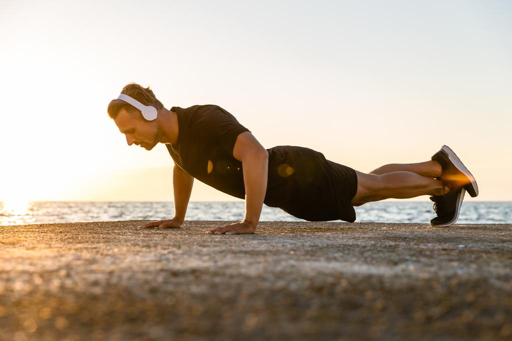 Strong adult sportsman in headphones doing push ups on seashore