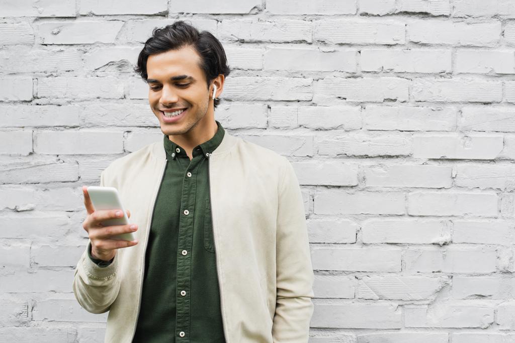 man in wireless earphones smiling while using smartphone near brick wall 