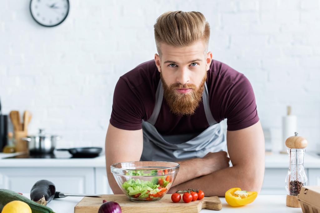 man in apron cooking vegetable salad and looking at camera