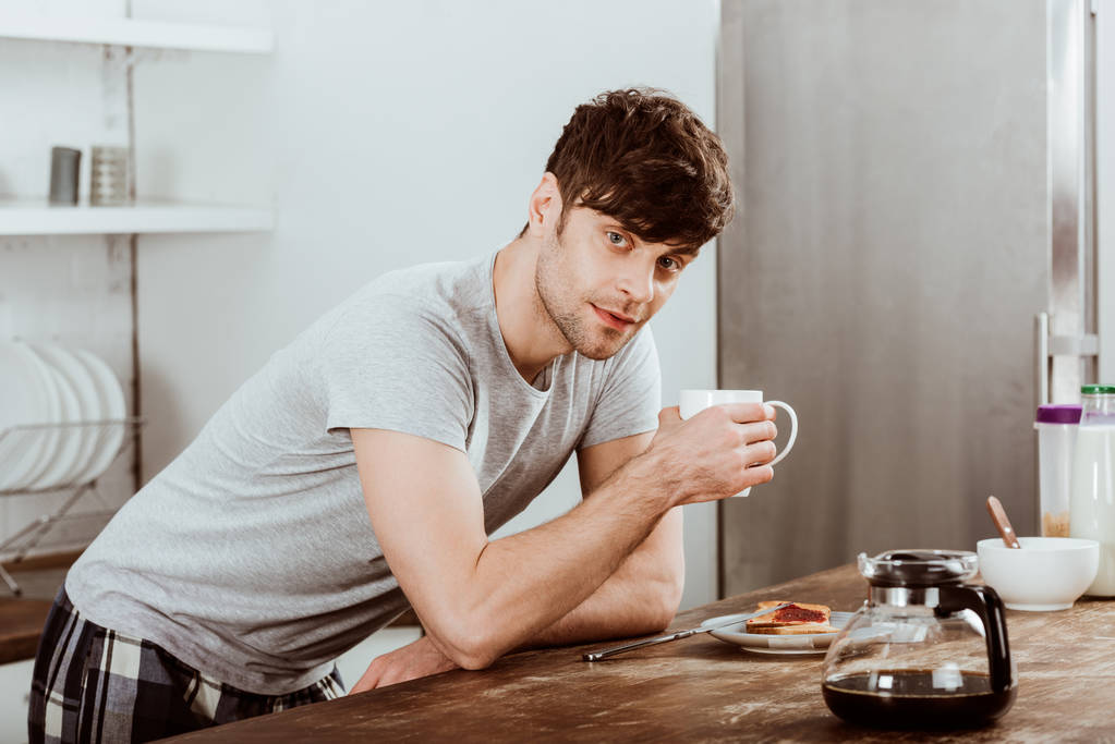 Man drinking coffee at table with toasts and coffee pot in kitchen at home