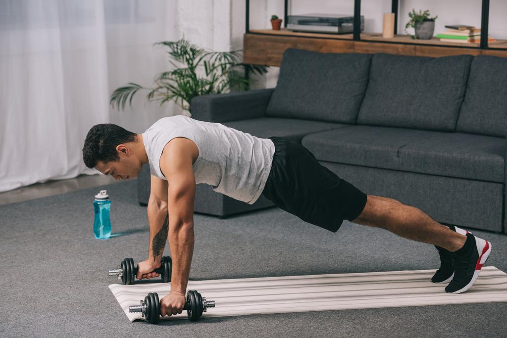 man doing plank exercise with dumbbells on fitness mat in living room