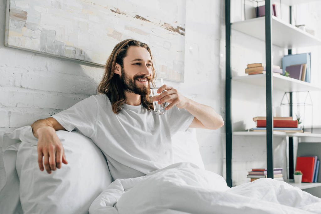 Smiling bearded man with long hair drinking water in bedroom at home