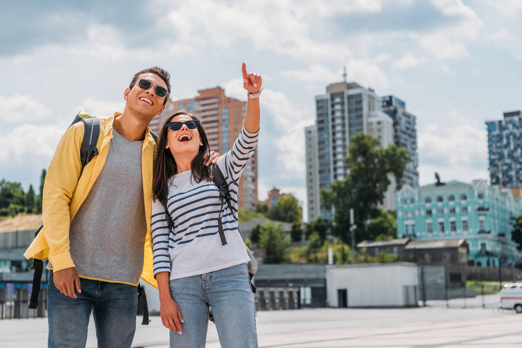 Woman pointing with finger and smiling near bi-racial friend with backpack