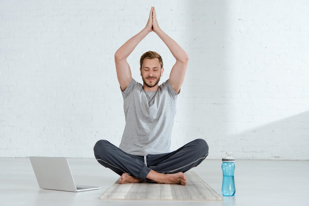  man practicing half lotus pose with raised prayer hands near laptop and sports bottle