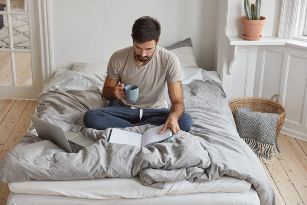 young caucasian man has morning coffee, sits crossed legs on bed