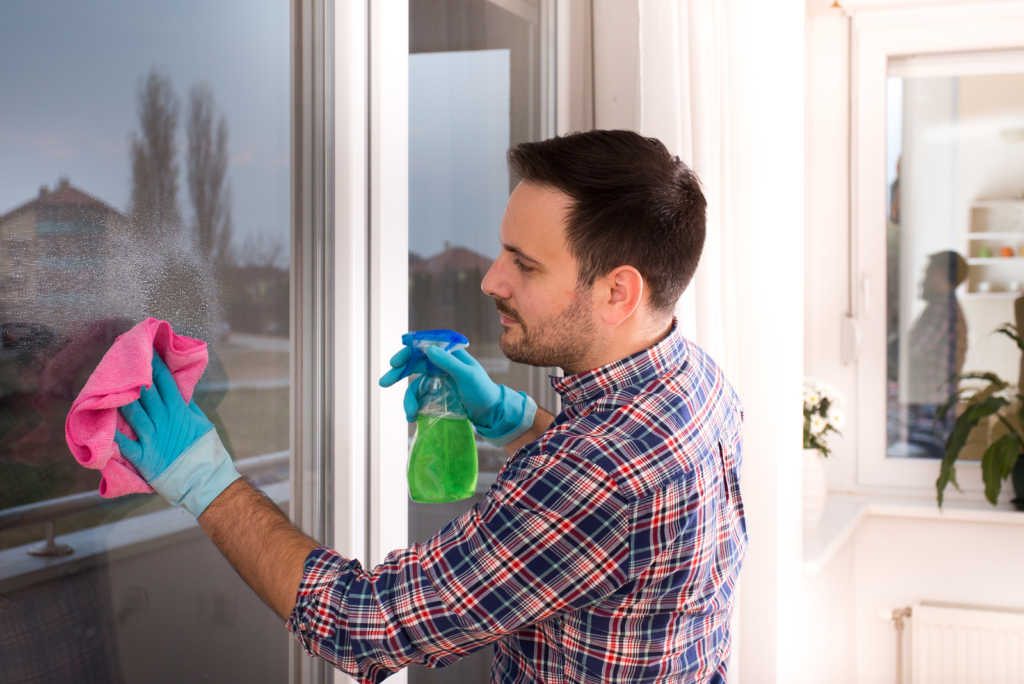 Handsome young man cleaning windows with cloth and spray bottle at home