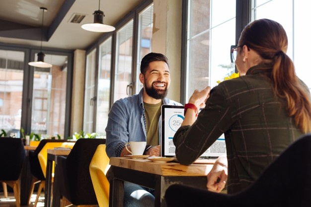 charming young man sitting in the cafe and smiling happily while working on the laptop