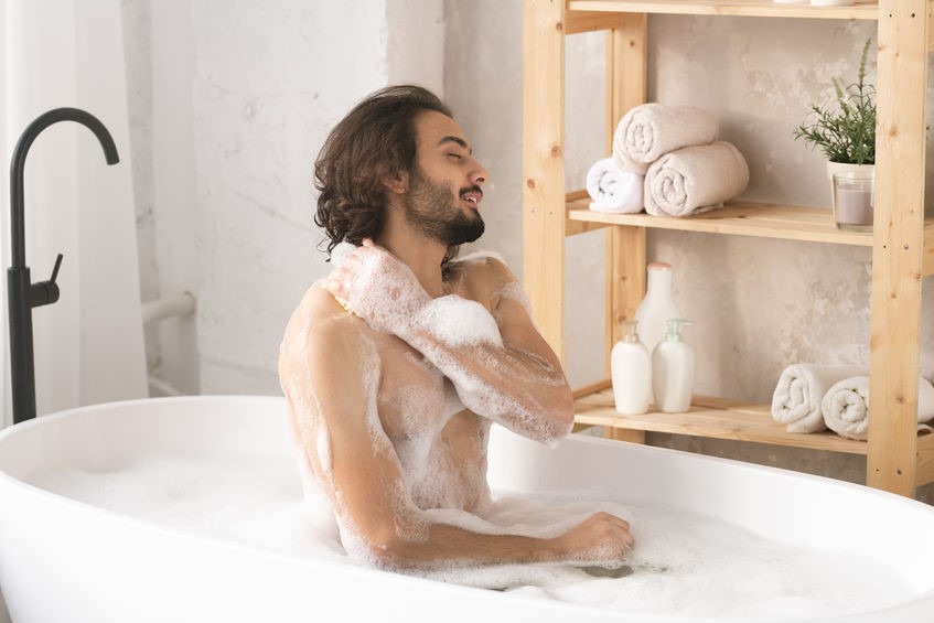 Young handsome naked man sitting in bath with hot water and foam, washing his body and enjoying the procedure 
