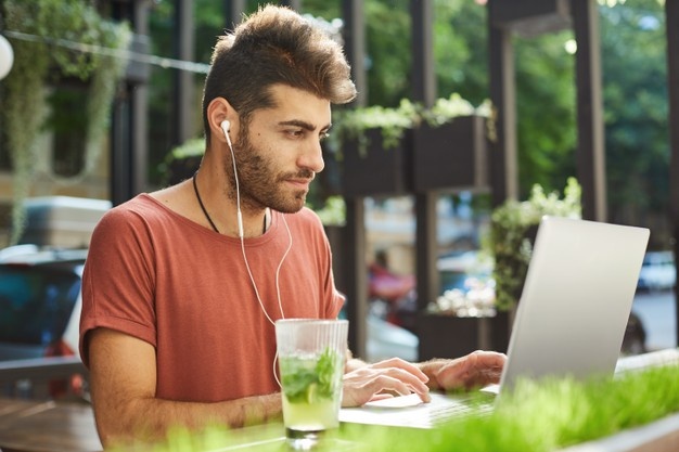 Handsome bearded man, freelancer working remote from outdoor cafe, programmer with laptop listening music to focus on work 